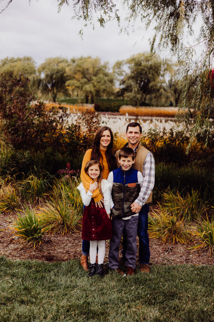 Family of four smiling together outdoors in Sheboygan, WI with autumn colors in the background during fall family session.