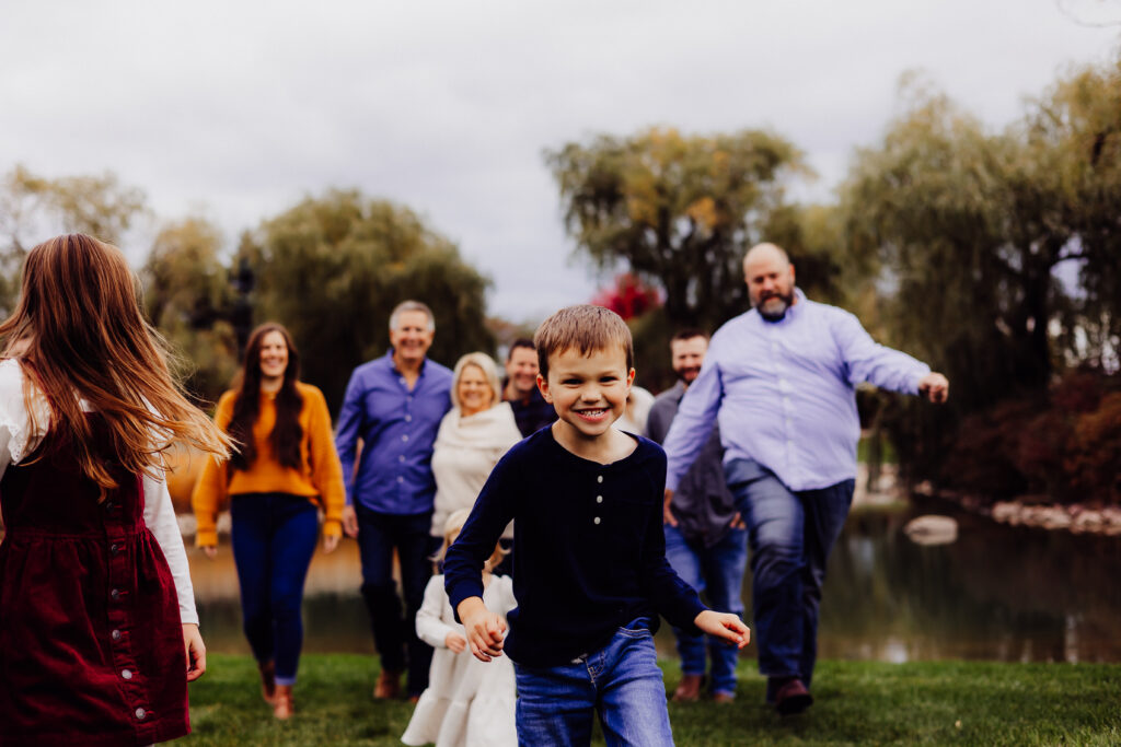 Smiling young boy walking toward the camera with extended family blurred in the background during autumn family session in Sheboygan, WI.