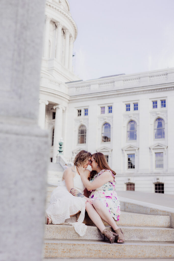 Lauren and Carley leaning close and smiling on the granite stairs of the Wisconsin State Capitol in Madison during their spring engagement session.