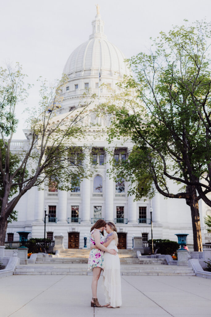 Lauren and Carley standing forehead to forehead with the Wisconsin State Capitol dome in the background during their romantic spring engagement photos.