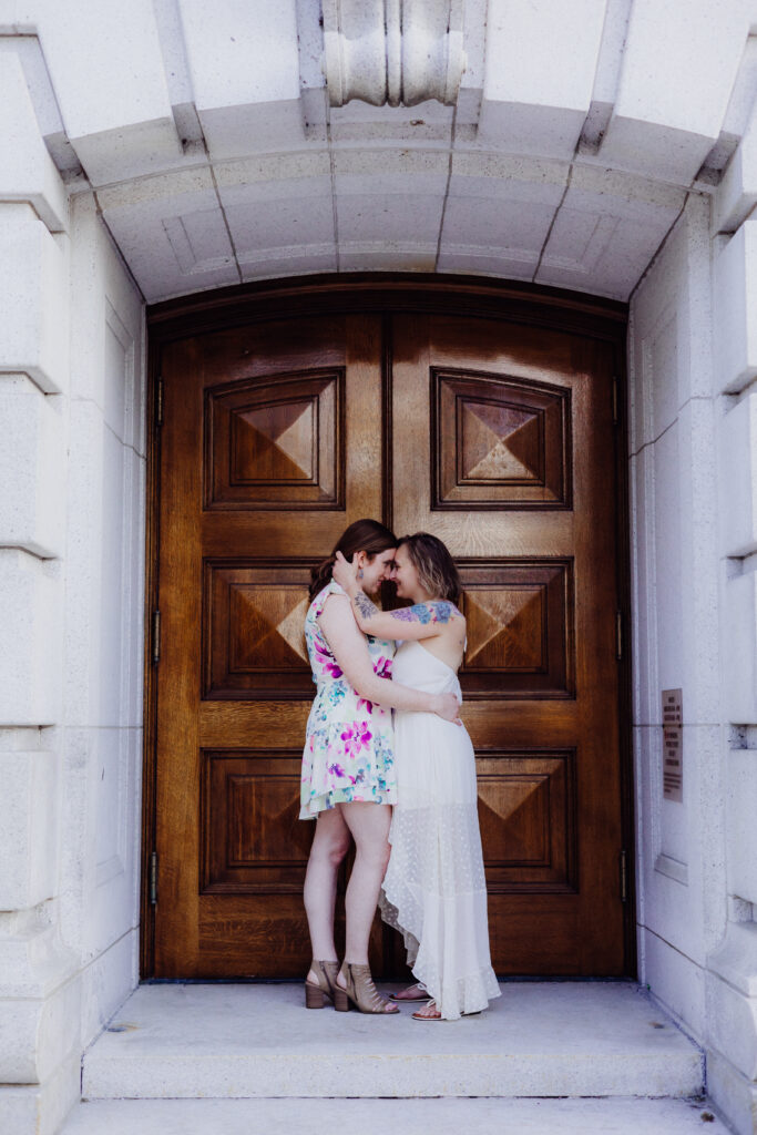 Lauren and Carley standing forehead to forehead, smiling softly as the sun sets behind them during their spring engagement session in downtown Madison, WI, wearing a white dress and a floral dress.