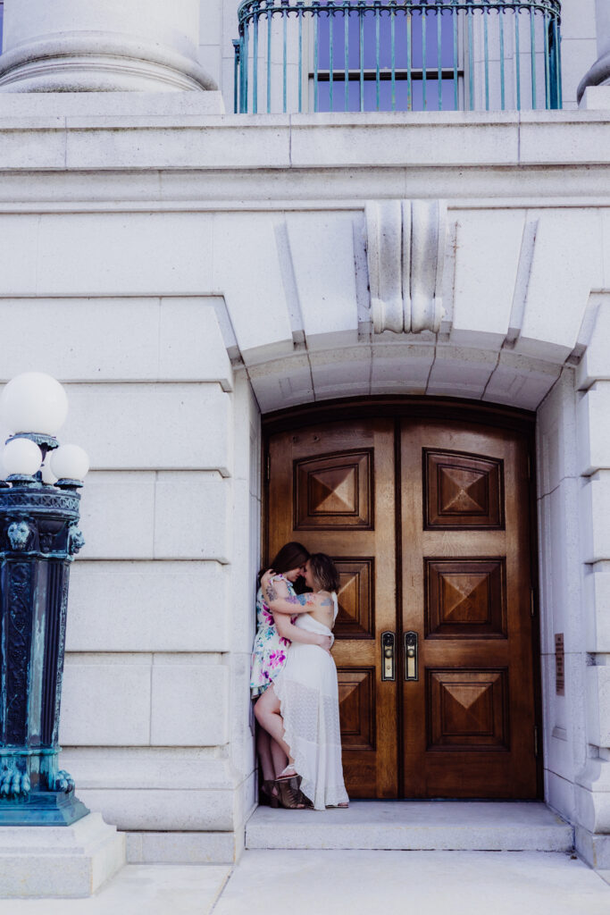 Lauren and Carley embracing closely in front of the large wooden doors of the Wisconsin State Capitol during their romantic spring engagement session in Madison, WI.