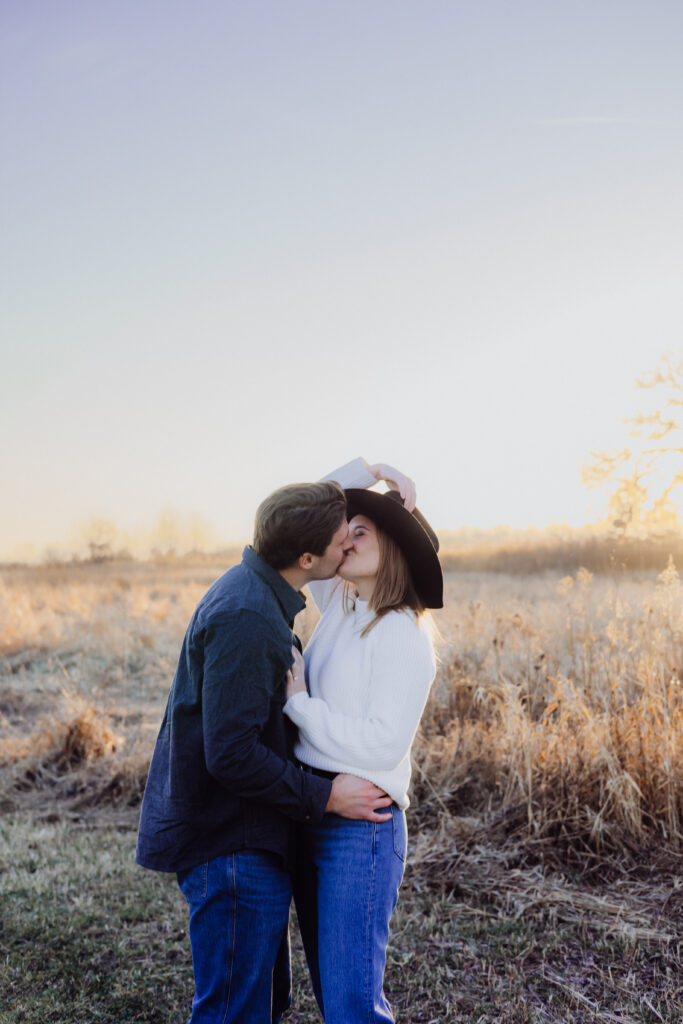 A couple shares a romantic kiss in a golden field at sunset. The woman, wearing a wide-brimmed black hat and white sweater, gently holds her partner while he wraps an arm around her waist. Warm sunlight creates a dreamy glow in the background.
