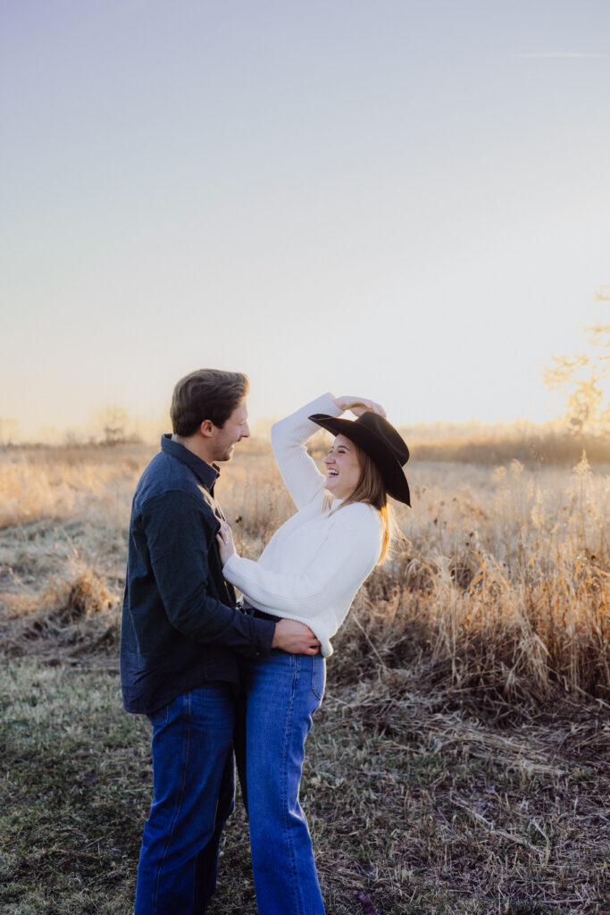A couple laughs together in a golden field at sunset. The woman, wearing a black hat and white sweater, tilts her head back with a smile as her partner holds her close. The warm sunlight glows through the tall grasses, creating a romantic, playful atmosphere.