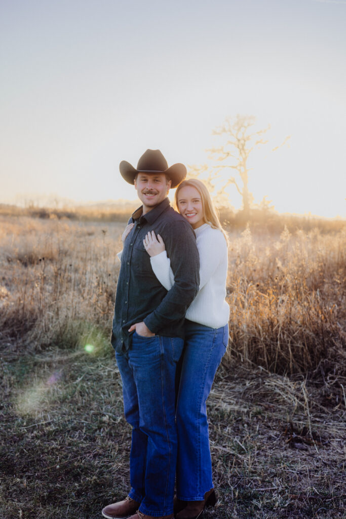 A couple poses in a golden field at sunset, with the man in a cowboy hat and dark shirt standing in front and smiling at the camera. The woman, in a white sweater and jeans, embraces him from behind with a bright smile as sunlight glows warmly through the tall grass.