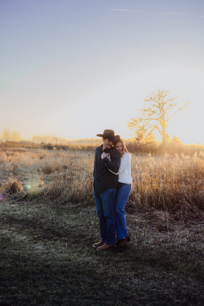 A couple embraces in a golden field at sunset. The man, wearing a cowboy hat and dark shirt, smiles softly while looking down, as the woman in a white sweater wraps her arms around him from behind. The tall grasses and glowing sun create a warm, romantic country atmosphere.