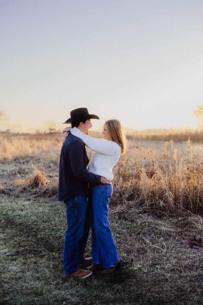 A couple embraces in a golden field at sunset. The man, wearing a cowboy hat and dark shirt, smiles at his partner while holding her waist. The woman, in a white sweater and jeans, wraps her arms around his neck, both sharing a joyful gaze as the evening light glows behind them.