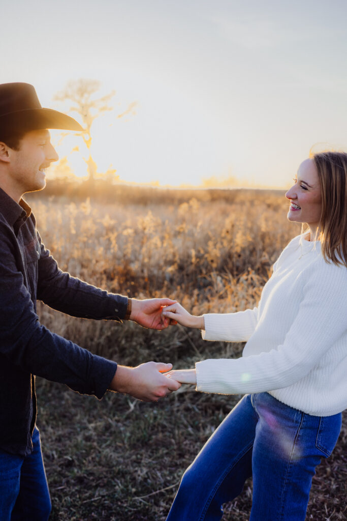 A couple holds hands and smiles at each other in a golden field during sunset. The man, wearing a cowboy hat and dark shirt, faces his partner while she leans back slightly, laughing in a white sweater and jeans. The warm light creates a playful and romantic atmosphere.
