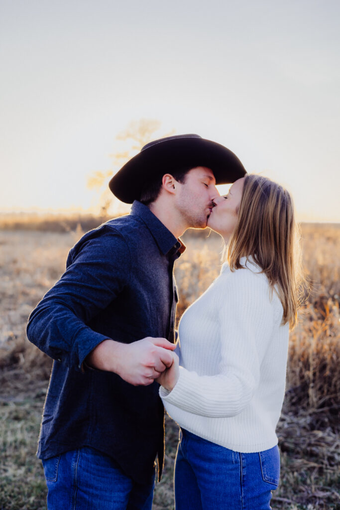 A couple kisses in a golden field at sunset. The man, wearing a cowboy hat and dark shirt, holds his partner’s hand as she leans in close in a white sweater and jeans. The warm light creates a soft, romantic glow around them.