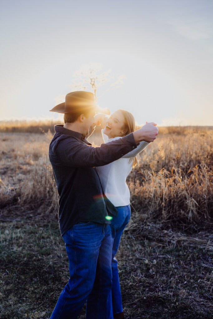 A couple dances playfully in a golden field at sunset. The man, wearing a cowboy hat and dark shirt, holds his partner’s hands while she laughs joyfully in a white sweater and jeans. Sunlight streams between them, casting a warm, romantic glow.