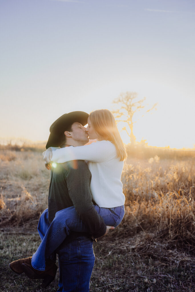 A couple kisses in a golden field at sunset. The man, wearing a cowboy hat and dark shirt, lifts his partner in his arms as she wraps her arms around his shoulders. The woman, dressed in a white sweater and jeans, leans in close with the sun glowing warmly behind them.