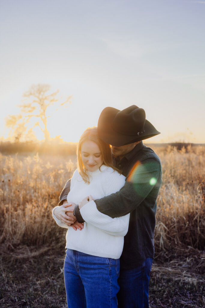 A couple shares a quiet embrace in a golden field at sunset. The man, wearing a cowboy hat, wraps his arms around his partner from behind as she smiles softly and looks down. The warm light and lens flare create a tender, romantic atmosphere.
