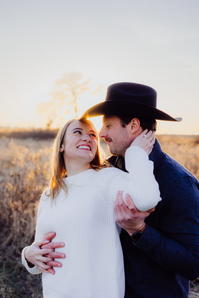A couple stands close together in a golden field at sunset. The woman, smiling brightly in a white sweater, looks up at her partner while resting her hand on his neck. The man, wearing a cowboy hat and dark shirt, gazes back at her with the sun glowing warmly behind them.