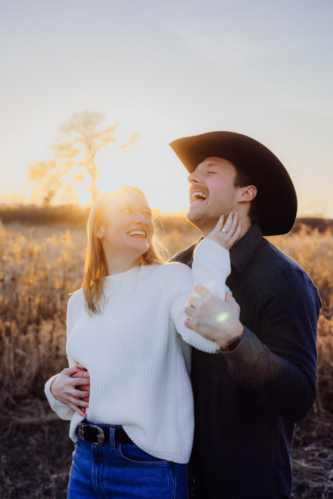 A couple laughs together in a golden field at sunset. The woman, in a white sweater, smiles up at her partner while resting her hand on his neck. The man, wearing a cowboy hat and dark shirt, throws his head back in laughter as the sun glows warmly behind them.