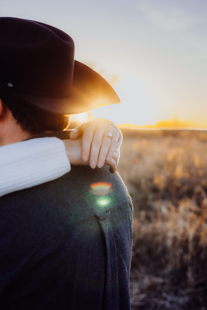 Close-up of a woman’s hand resting on her partner’s shoulder, showing her engagement ring as the sun sets behind them. The man wears a cowboy hat and dark shirt, with warm golden light and lens flare creating a romantic glow in the background.