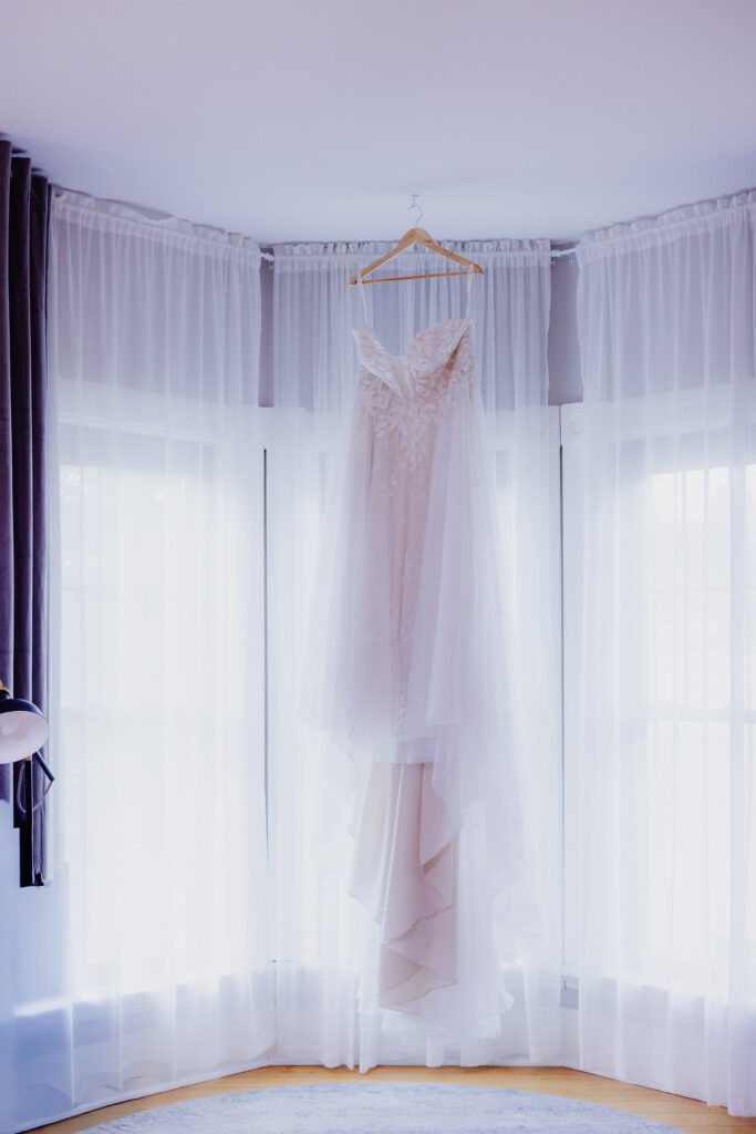A delicate white lace wedding gown hangs gracefully on a wooden hanger in front of sheer floor-to-ceiling curtains. Soft natural light pours through the windows, highlighting the dress’s floral details and flowing layers, creating a romantic and timeless bridal scene.