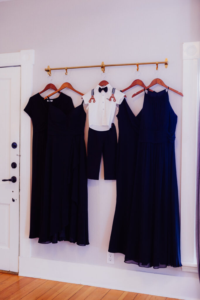 Three elegant black bridesmaid dresses and a toddler’s white shirt with suspenders, bow tie, and black pants hang neatly on wooden hangers along a gold bar against a soft beige wall. The coordinated outfits are displayed together, ready for a wedding celebration.