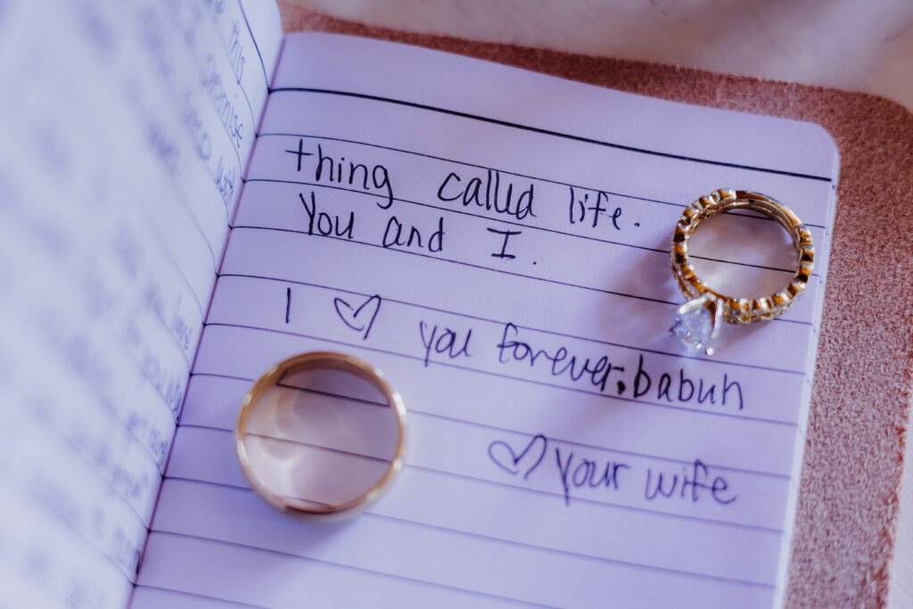 A pair of wedding rings rest on the open pages of a handwritten love note. The gold band and diamond engagement ring are placed above words that read, “I ♥ you forever, babuh. ♥ Your wife,” symbolizing love and commitment on a wedding day.