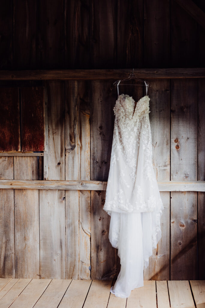 Lace off-the-shoulder wedding gown with floral appliqués hanging on rustic wooden barn wall before the ceremony in Juneau, Wisconsin.