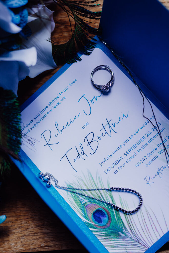Wedding invitation with teal lettering and peacock feather accents surrounded by engagement ring, necklace, and jewelry, styled on wood