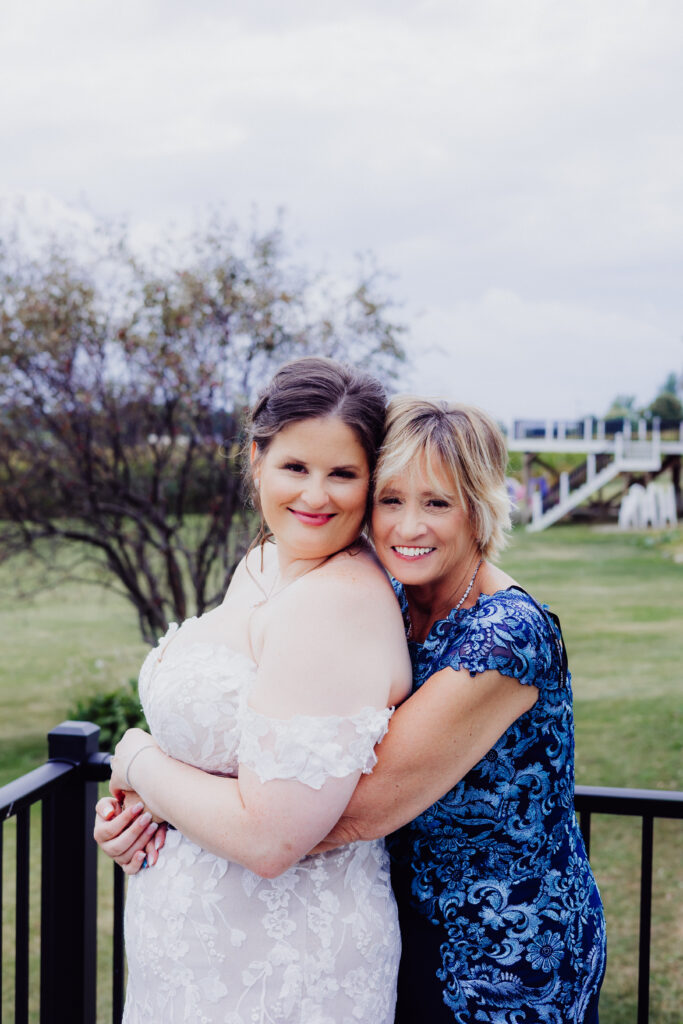 Bride in lace gown embraces her mother in a navy floral dress outdoors before barn wedding ceremony in Juneau, Wisconsin.