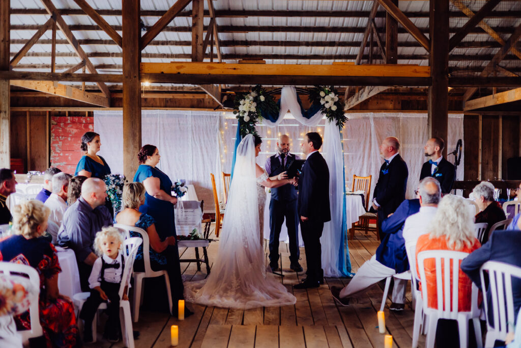 Bride and groom exchange vows under a floral-draped arch inside rustic barn, surrounded by bridesmaids and groomsmen, with family looking on