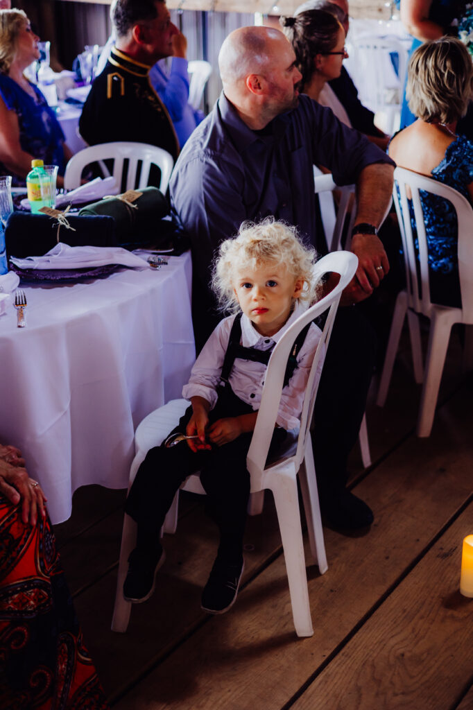 Young boy with curly blonde hair sits in a white chair at a rustic barn wedding reception in Juneau, Wisconsin, wearing a white shirt and black suspenders.
