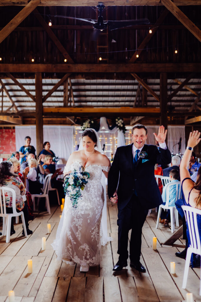 Newlywed couple smiles and walks hand in hand down the barn aisle as guests clap, bride holding blue floral bouquet and groom waving.