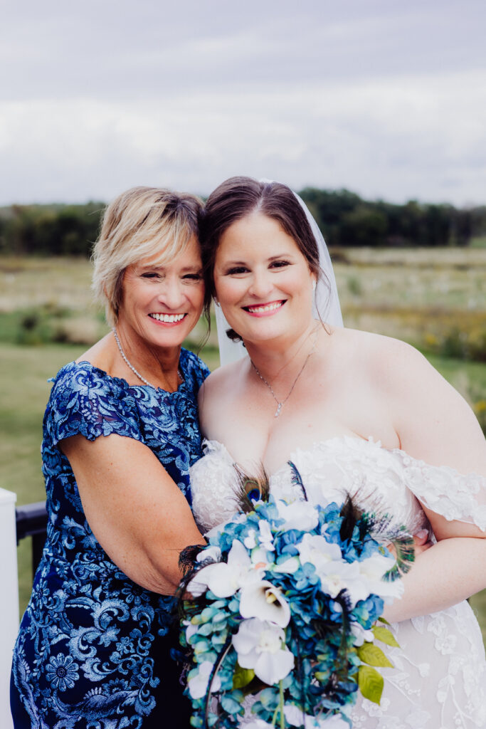 Bride holding blue and white bouquet stands smiling with her mother, both in lace dresses, outdoors at barn wedding.