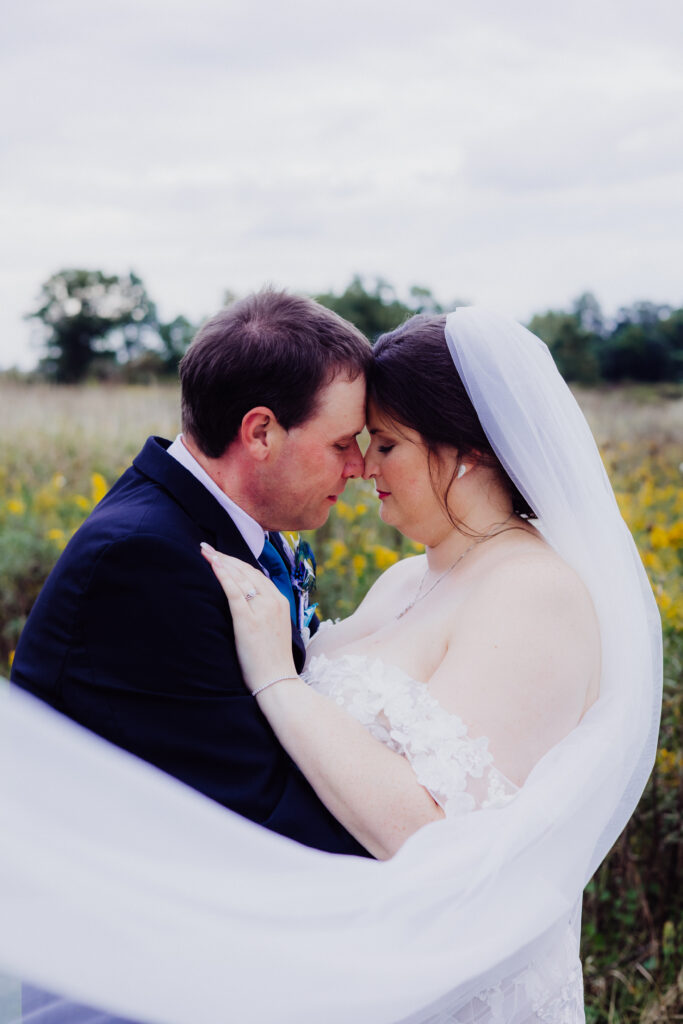 Groom embraces bride from behind in field, both laughing and smiling during romantic countryside wedding portraits.