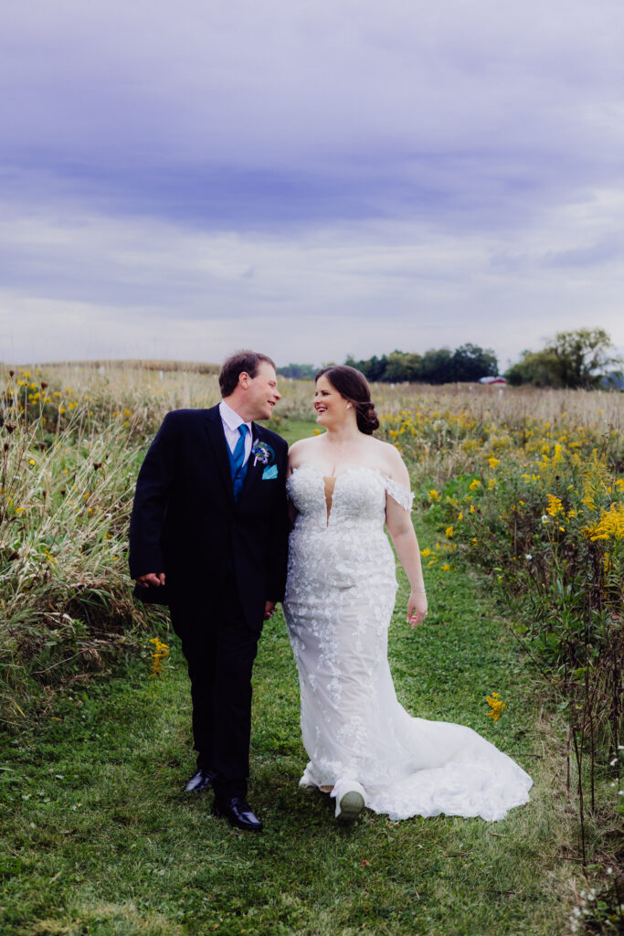 Bride and groom walk hand in hand along grassy path surrounded by tall wildflowers and countryside fields under moody Wisconsin sky.